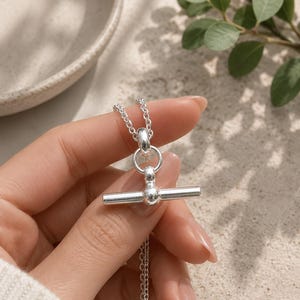 May include: A shiny silver necklace featuring a toggle clasp with a horizontal bar and a circular ring. The necklace is constructed from a delicate chain with small, uniform links that connect to the central toggle mechanism, creating a clean look.