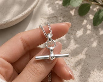 May include: A shiny silver necklace featuring a toggle clasp with a horizontal bar and a circular ring. The necklace is constructed from a delicate chain with small, uniform links that connect to the central toggle mechanism, creating a clean look.