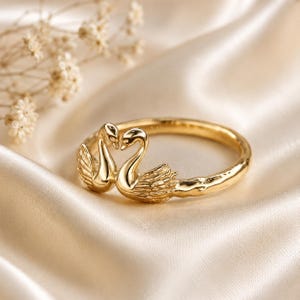 May include: A gold ring featuring two sculpted swans facing each other with their necks curved to form a heart shape. The band has a textured, organic finish and is stamped with 14k on the interior surface of the metal ring band.