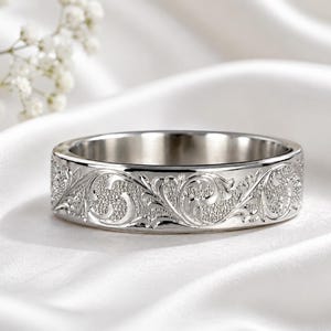 May include: A silver-toned metal ring featuring a flat band with an intricate, raised scrollwork pattern engraved along the exterior surface. The design includes detailed swirling floral motifs set against a textured, matte background finish.