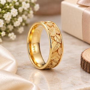 May include: A gold-toned ring featuring an intricate, embossed pattern of overlapping leaves around the exterior band. The interior surface of the ring is smooth and polished, clearly displaying the word Love engraved in a clean, sans-serif font.