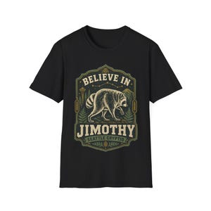 以下が含まれることがあります： 森の中にいるアライグマのグラフィックプリントが施された黒の半袖Tシャツ。テキストには「BELIEVE IN JIMOTHY SEATTLE CRYPTID」とあり、スペースニードル、山、シダ、松ぼっくりのイラストが装飾的なフレームの中に描かれています。