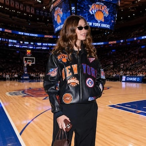 May include: A black leather varsity jacket featuring multiple New York Knicks basketball team patches, including the team name in orange and blue lettering, paired with black trousers and small dark-tinted sunglasses in a basketball arena.