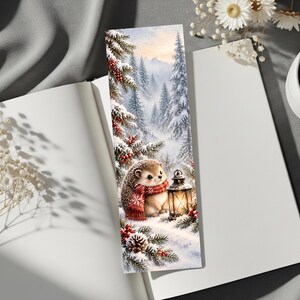 May include: A rectangular bookmark featuring a winter forest scene with a small hedgehog wearing a red patterned scarf. The hedgehog stands next to a glowing lantern in the snow, surrounded by pine trees, red berries, and pinecones.