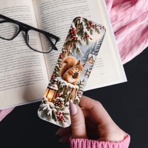 May include: A rectangular bookmark featuring a winter scene with a brown squirrel holding a pinecone, a glowing lantern, snow-covered pine branches with red berries, and a distant cabin, printed on a smooth surface with rounded corners.