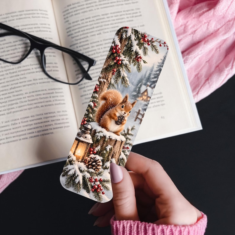 May include: A rectangular bookmark featuring a winter scene with a brown squirrel holding a pinecone, a glowing lantern, snow-covered pine branches with red berries, and a distant cabin, printed on a smooth surface with rounded corners.