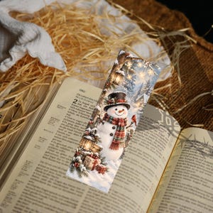 May include: A rectangular bookmark featuring a printed illustration of a snowman wearing a black top hat and a red and green plaid scarf. The scene includes a lantern, pine branches, wrapped gifts, and a small red bird in a snowy forest setting.
