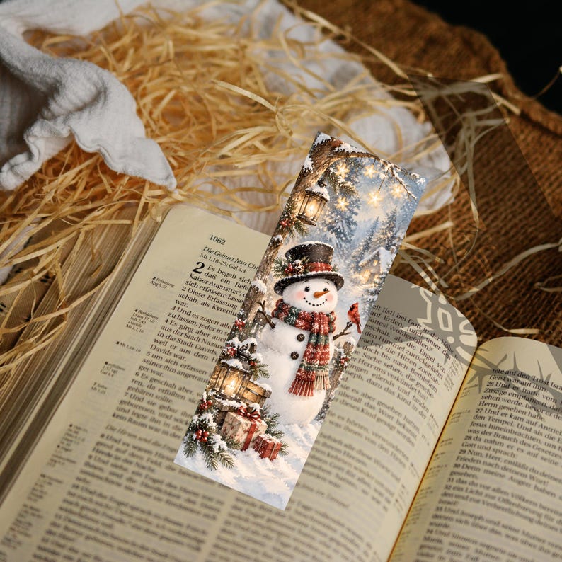 May include: A rectangular bookmark featuring a printed illustration of a snowman wearing a black top hat and a red and green plaid scarf. The scene includes a lantern, pine branches, wrapped gifts, and a small red bird in a snowy forest setting.