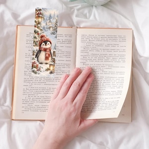 May include: A rectangular paper bookmark featuring a winter scene with a penguin wearing a red knit hat and scarf. The illustration includes snow-covered pine trees, glowing lanterns, and gift boxes. The bookmark is placed inside an open book.