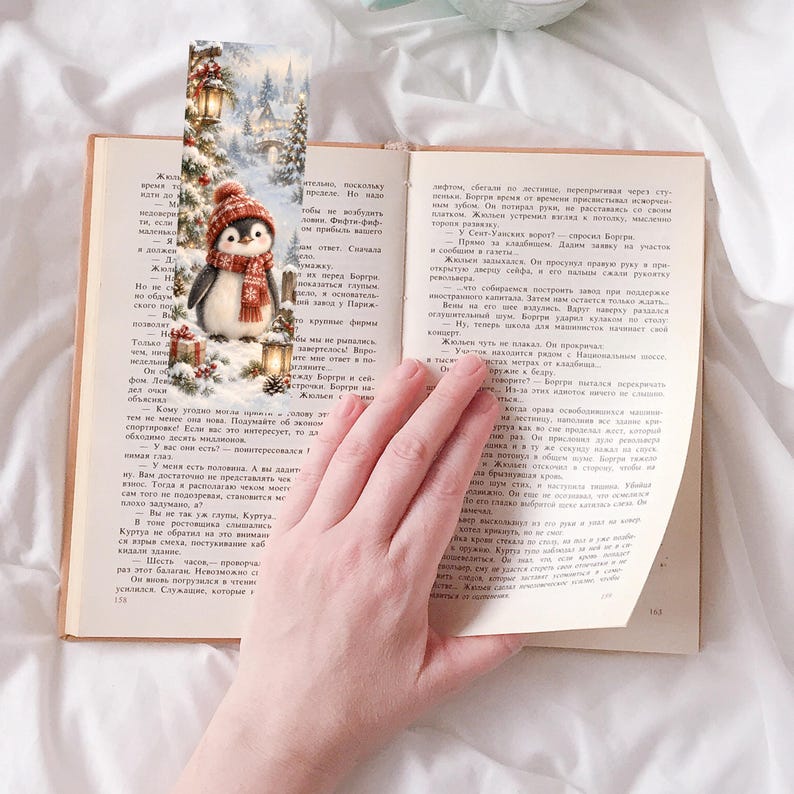 May include: A rectangular paper bookmark featuring a winter scene with a penguin wearing a red knit hat and scarf. The illustration includes snow-covered pine trees, glowing lanterns, and gift boxes. The bookmark is placed inside an open book.