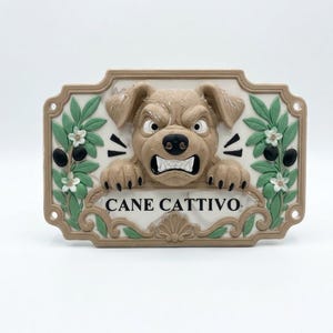 Può includere: Una targa da parete rettangolare raffigurante un cane color marrone chiaro scolpito con un'espressione arrabbiata, che mostra i denti. La targa è decorata con rami d'ulivo verdi e fiori bianchi su entrambi i lati, con la scritta CANE CATTIVO in nero.