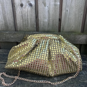 Op de afbeelding: Een gouden clutch met lovertjes, een geplooide, buidelachtige vorm en een dunne, metalen schouderketting. De buitenkant is bedekt met kleine, reflecterende gouden lovertjes die zorgen voor een getextureerd en glinsterend uiterlijk.