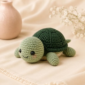 May include: A small, handmade crochet turtle toy featuring a light green head with black bead eyes and pink stitched cheeks. The shell is a dark forest green with a textured pattern, and the underside is a light beige color with four flippers.