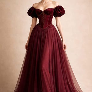 May include: A floor-length burgundy gown featuring a velvet corset-style bodice with a sweetheart neckline, short puffed velvet sleeves, and a voluminous tulle skirt. The bodice includes vertical boning details and a central front closure.