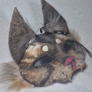 Könnte beinhalten: Eine katzenartige Maske mit einer strukturierten, gefleckten braun-grauen fellartigen Oberfläche und spitzen Ohren. Die Maske verfügt über schwarze Netz-Augenabdeckungen, eine rosa Nase, weiße Akzente über den Augen sowie dekorative schwarze Kordeln.