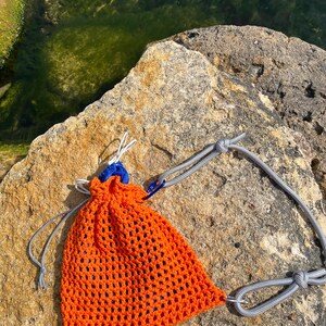 May include: A bright orange crocheted drawstring pouch featuring a wide-gauge mesh pattern. The bag includes a blue plastic cord lock, white drawstring cords, and a grey rope strap attached with a blue metal carabiner clip for carrying.