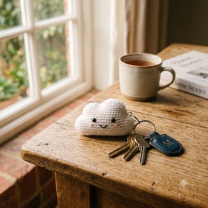 May include: A white crocheted cloud keychain with black bead eyes, a stitched smile, and pink cheeks. The cloud is attached to a metal keyring holding several silver keys and a small, dark blue leather tag embossed with the letter M in gold.