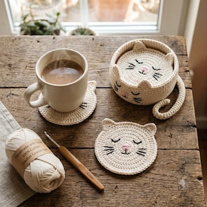 May include: A set of cream-colored crochet cat coasters, one placed under a white ceramic mug, one resting outside a matching cat-shaped holder, and a ball of cotton yarn with a 4.0mm bamboo crochet hook labeled Handmade with Love 100% Cotton.