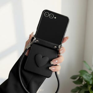 Puede incluir: Una funda de smartphone negra con acabado mate, que presenta un emblema en forma de corazón en relieve en la parte posterior. La funda incluye herrajes de metal plateado para una correa larga negra, permitiendo llevar el dispositivo cruzado.