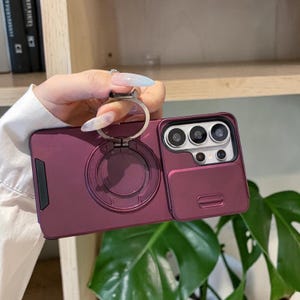 Puede incluir: Una funda de smartphone de color morado oscuro con un soporte de anillo circular de metal integrado en la parte posterior y una cubierta deslizante para la lente de la cámara. La funda tiene un acabado mate con un detalle negro lateral.