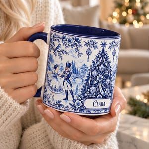 May include: A white ceramic mug with a dark blue interior and handle, featuring a detailed blue and white pattern of a nutcracker, a ballerina, and a decorated Christmas tree. The name Clara is printed in a rectangular frame at the bottom.