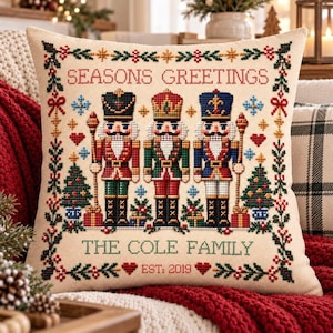 May include: A square throw pillow with a cream-colored fabric featuring a cross-stitched design of three nutcrackers in red, green, and blue uniforms. The text reads SEASONS GREETINGS, THE COLE FAMILY, and EST: 2019, framed by a festive holiday border.