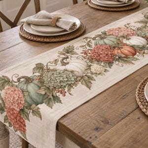May include: A rectangular table runner featuring a printed pattern of pumpkins, hydrangea flowers, and berries in muted shades of green, orange, and cream. The fabric has a textured, linen-like appearance with a neutral beige background color.