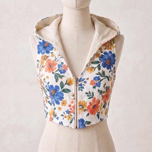 May include: A sleeveless, cropped vest featuring a hood and a front zipper closure. The garment is made of white fabric printed with a floral pattern consisting of blue, orange, and yellow flowers with green leaves, and has a solid cream lining.