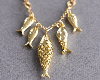 Puede incluir: Un collar de cadena dorada con cinco colgantes de peces dorados. Los colgantes de peces están detallados con escamas y tienen una pequeña piedra transparente en sus cuerpos.
