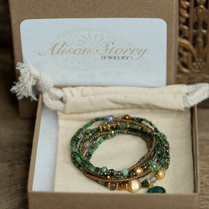 K&ouml;nnte beinhalten: Ein gr&uuml;nes und goldenes Edelstein-Armband mit einem Anh&auml;nger, verpackt in einer braunen Schachtel mit einer wei&szlig;en Karte, auf der "Alison Story Jewelry" steht.