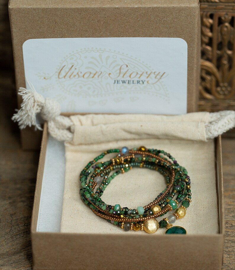 K&ouml;nnte beinhalten: Ein gr&uuml;nes und goldenes Edelstein-Armband mit einem Anh&auml;nger, verpackt in einer braunen Schachtel mit einer wei&szlig;en Karte, auf der "Alison Story Jewelry" steht.