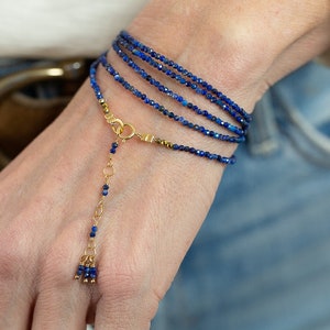Puede incluir: Un brazalete de múltiples hilos hecho de pequeñas cuentas de lapislázuli azul oscuro. El brazalete tiene un cierre dorado y un dije colgante con un borla de cuentas de lapislázuli.
