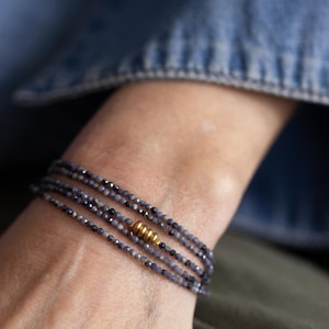 May include: A multi-strand bracelet with small, dark blue beads and a gold-colored accent. The bracelet is wrapped around a wrist, with a denim jacket in the background. The beads appear to be approximately 0.1 inches in diameter.