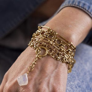 May include: A gold chain bracelet with multiple layers of different chain styles. The bracelet has a large, round, open-link charm and a small, white, teardrop-shaped charm.
