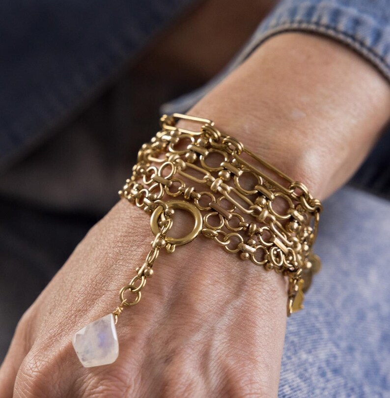 May include: A gold chain bracelet with multiple layers of different chain styles. The bracelet has a large, round, open-link charm and a small, white, teardrop-shaped charm.