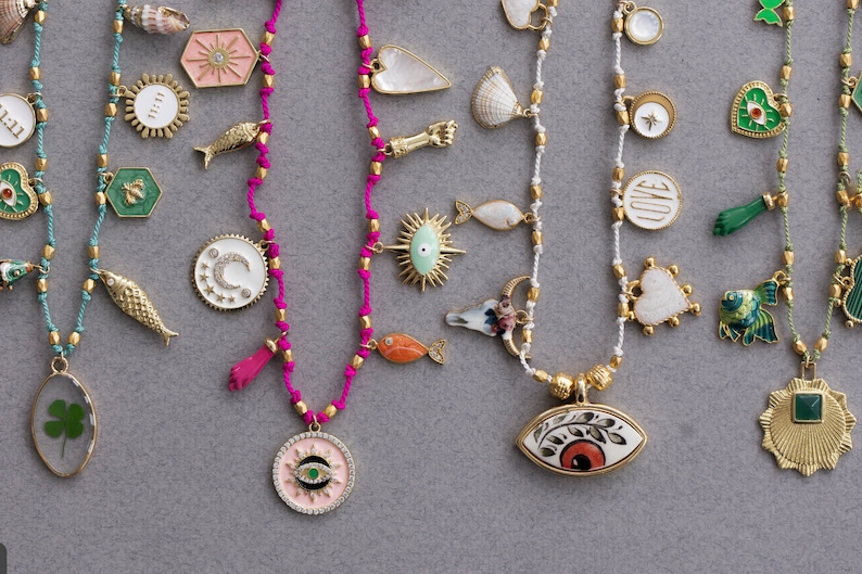 May include: A collection of colorful necklaces with various charms, including fish, hearts, stars, moons, and eyes. The necklaces are made with different colored cords and beads.