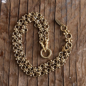 Könnte beinhalten: Eine goldfarbene Halskette mit einem einzigartigen Kettendesign. Die Halskette besteht aus einer Reihe kleiner, runder Perlen und ineinandergreifenden Gliedern, wodurch ein mehrschichtiger Effekt entsteht. Sie hat einen großen kreisförmigen Verschluss und einen kleineren Verschluss zum Schließen. Die Halskette wird auf einer Holzoberfläche präsentiert.