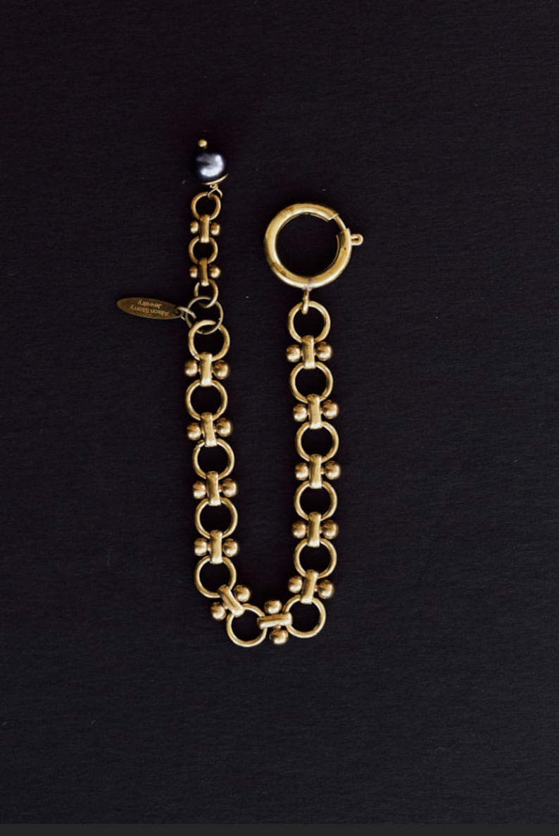 Puede incluir: Una cadena dorada con un cierre y una peque&ntilde;a perla oscura. La cadena est&aacute; compuesta por anillos interconectados y elementos decorativos. Una peque&ntilde;a etiqueta con texto est&aacute; adjunta. El fondo es negro.