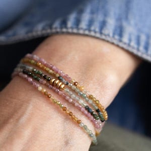 Pu&ograve; includere: Un braccialetto a pi&ugrave; fili con piccole perline di pietre preziose colorate nelle tonalit&agrave; del rosa, verde, giallo e marrone. Il braccialetto ha una chiusura color oro e viene indossato al polso. Lo sfondo &egrave; una camicia di jeans sfocata.