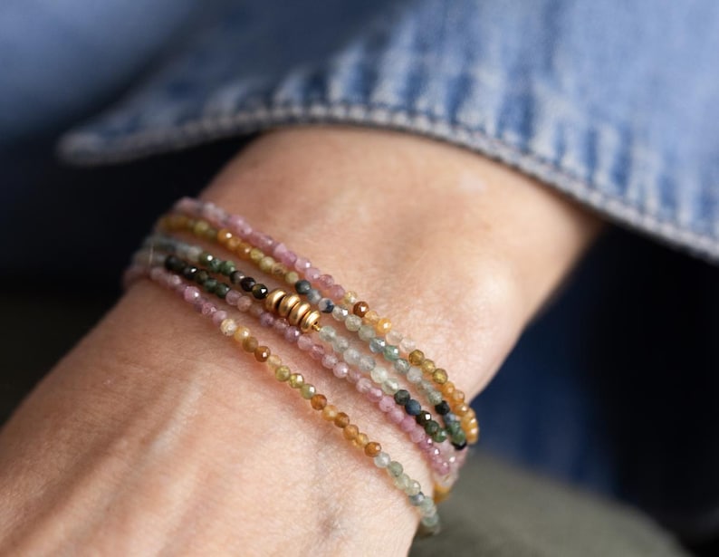 Pu&ograve; includere: Un braccialetto a pi&ugrave; fili con piccole perline di pietre preziose colorate nelle tonalit&agrave; del rosa, verde, giallo e marrone. Il braccialetto ha una chiusura color oro e viene indossato al polso. Lo sfondo &egrave; una camicia di jeans sfocata.