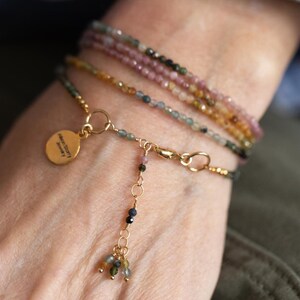 Pu&ograve; includere: Una collezione di delicati braccialetti di perline nei toni del rosa, verde e oro. Un braccialetto presenta un ciondolo dorato con testo e un gruppo pendente di perline colorate. I braccialetti sono stratificati su un polso.