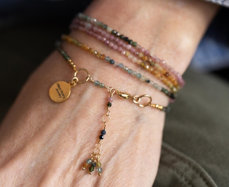 Pu&ograve; includere: Una collezione di delicati braccialetti di perline nei toni del rosa, verde e oro. Un braccialetto presenta un ciondolo dorato con testo e un gruppo pendente di perline colorate. I braccialetti sono stratificati su un polso.
