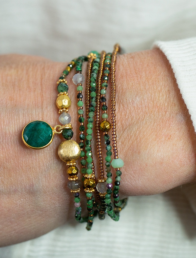 K&ouml;nnte beinhalten: Ein mehrreihiges Armband mit gr&uuml;nen, braunen und goldenen Perlen. Das Armband hat einen gro&szlig;en Anh&auml;nger aus gr&uuml;nem Edelstein.