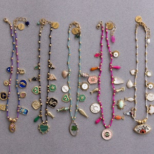 May include: Ten gold necklaces with charms, each featuring a different design. The charms include a heart, a four-leaf clover, a crescent moon, a sun, an eye, a fish, a seashell, a star, and a cross. The necklaces are made of gold beads and are strung on a thin gold chain.