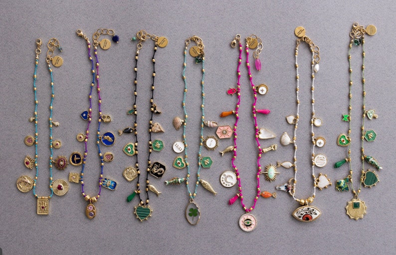 May include: Ten gold necklaces with charms, each featuring a different design. The charms include a heart, a four-leaf clover, a crescent moon, a sun, an eye, a fish, a seashell, a star, and a cross. The necklaces are made of gold beads and are strung on a thin gold chain.