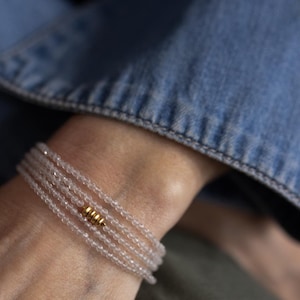 K&ouml;nnte beinhalten: Ein zartes Armband aus mehreren Str&auml;ngen kleiner, klarer Perlen. Ein goldfarbenes zylindrisches Akzentst&uuml;ck ist auf dem Armband zentriert. Das Armband wird an einem Handgelenk getragen, mit einer Jeansjacke im Hintergrund.