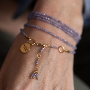 May include: A delicate bracelet featuring multiple strands of small, light purple beads. The bracelet includes gold-colored chain accents and a small, round charm. The charm has text on it. The bracelet is wrapped around a wrist.