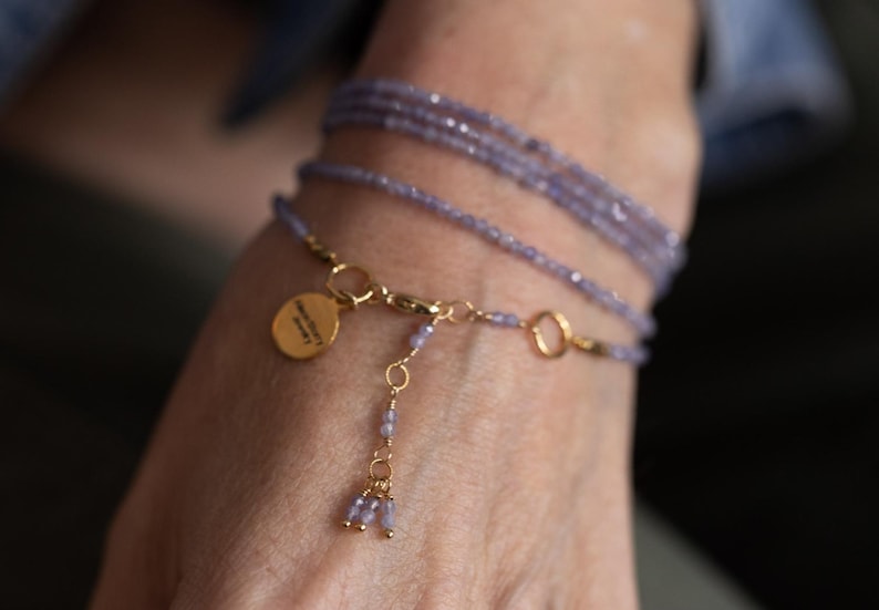 May include: A delicate bracelet featuring multiple strands of small, light purple beads. The bracelet includes gold-colored chain accents and a small, round charm. The charm has text on it. The bracelet is wrapped around a wrist.