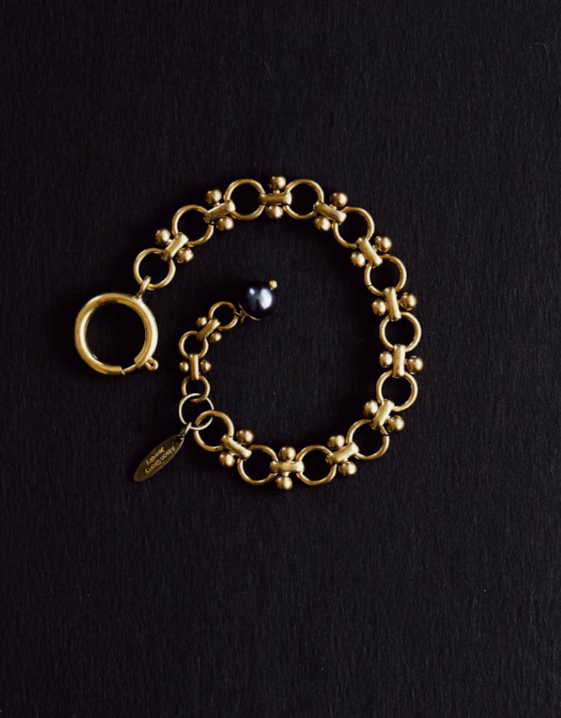 Puede incluir: Una pulsera dorada con un dise&ntilde;o de eslabones circulares y una peque&ntilde;a perla negra. La pulsera tiene un cierre de broche y una peque&ntilde;a etiqueta que dice "Alison Sherry Jewelry". La joya est&aacute; sobre un fondo negro.