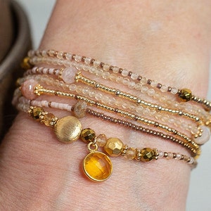 May include: A stack of delicate bracelets featuring a mix of gold, brown, and pink beads, with a gold charm and a yellow gemstone pendant.